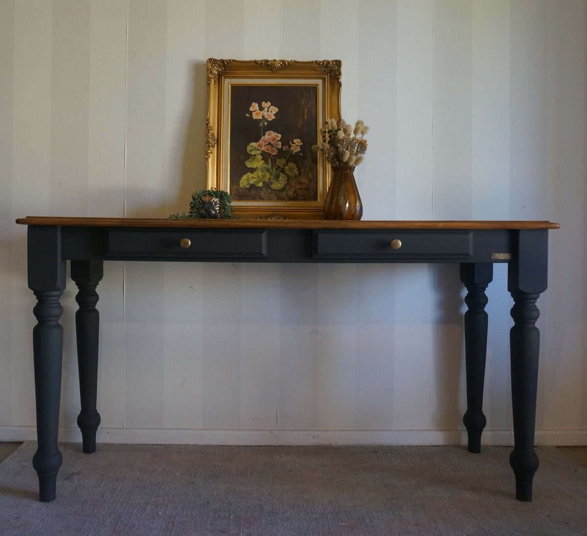 A dark black hall table with a walnut wooden top, featuring two drawers with brass knobs and turned legs. The table is styled with a framed floral painting and a vase of dried flowers against a white paneled wall.
