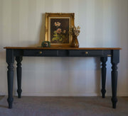 A dark black hall table with a walnut wooden top, featuring two drawers with brass knobs and turned legs. The table is styled with a framed floral painting and a vase of dried flowers against a white paneled wall.