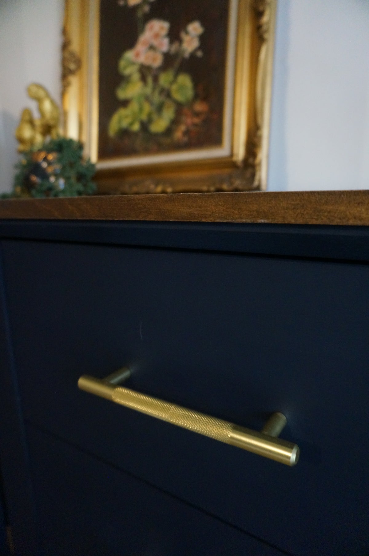A close-up of a dark navy bedside table featuring a long, vertical brushed gold metal handle.