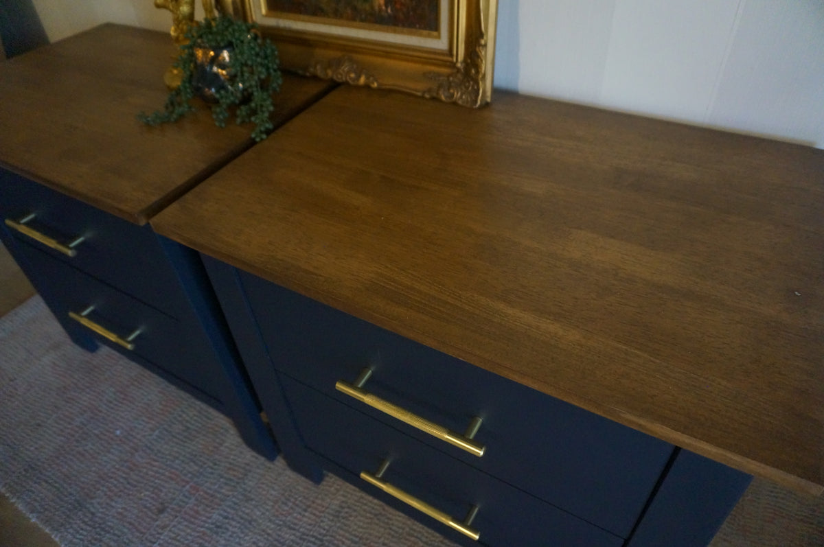 Dark blue bedside tables with two drawers and gold metal handles, shown in a home setting with a framed picture and greenery on top.