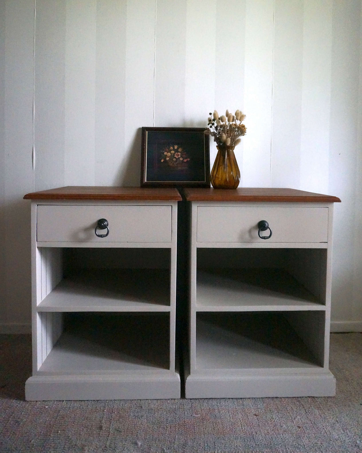 A pair of country style bedside tables with Velvet Taupe painted bodies, jarrah wood tops, and open shelving, set against a white paneled wall with decorative items on top.