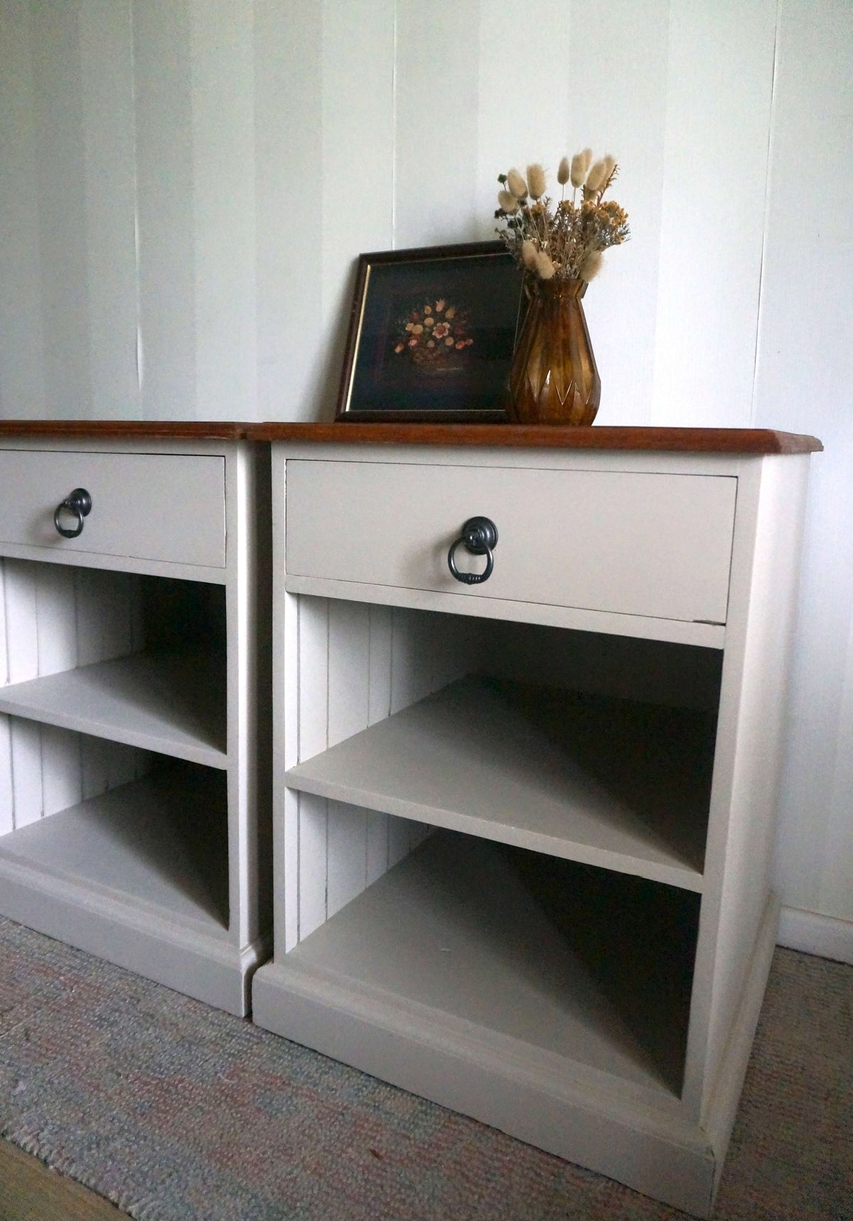 A pair of Velvet Taupe painted wooden bedside tables with a jarrah wood top, open shelving, and dark metal ring pulls, shown against a white paneled wall with decorative items on top.
