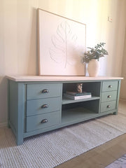 A kettle green pine TV unit with a light natural wood top, styled in a room setting with a large framed leaf print, a white vase with greenery, and books on the shelf.