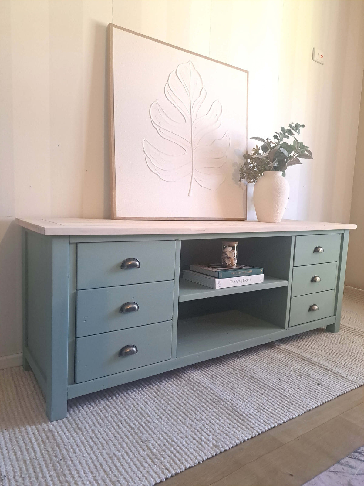 A kettle green pine TV unit with a light natural wood top, styled in a room setting with a large framed leaf print, a white vase with greenery, and books on the shelf.
