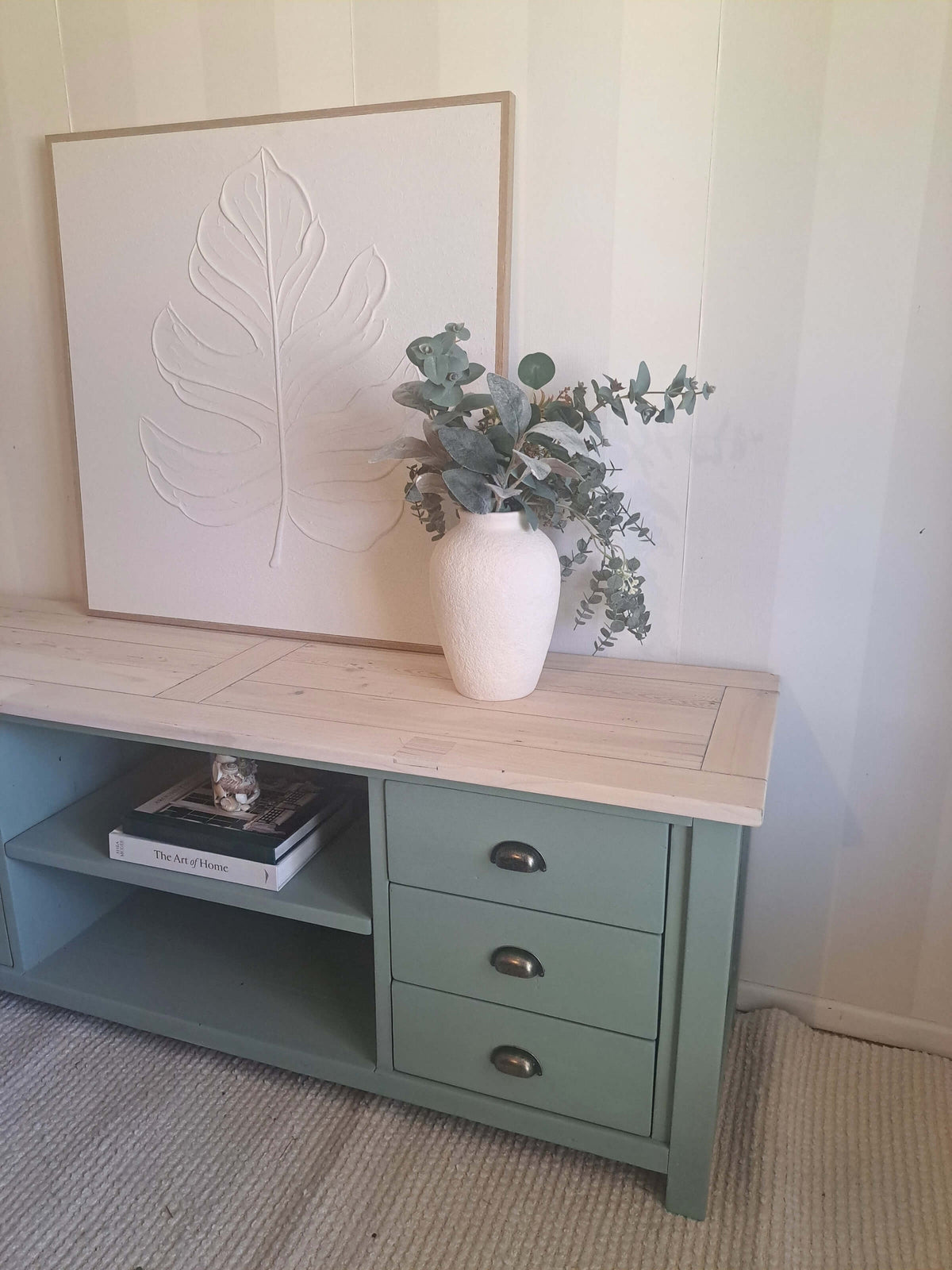 A kettle green pine TV unit TV with a light natural wood top, styled with a white ceramic vase of eucalyptus and a large framed leaf art print against a striped wall.