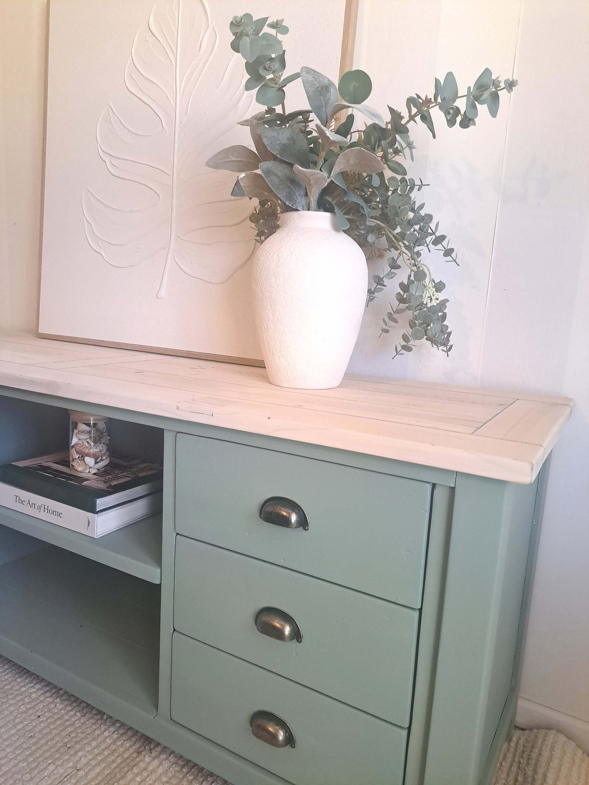 A kettle green painted TV unit with a light natural wood top, featuring three drawers with dark metal handles and an open shelf on the left. A white ceramic vase filled with eucalyptus branches sits on the table, set against a white paneled wall with a large leaf-shaped wall art piece.