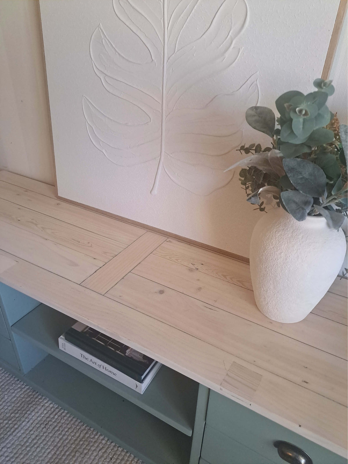 A light natural wood TV unit with a textured white ceramic vase of eucalyptus on top, set against a white wall with a large embossed leaf art piece.