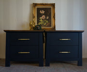 A pair of dark navy blue nightstands with gold metal handles, shown side-by-side against a white paneled wall with a framed floral painting on top.