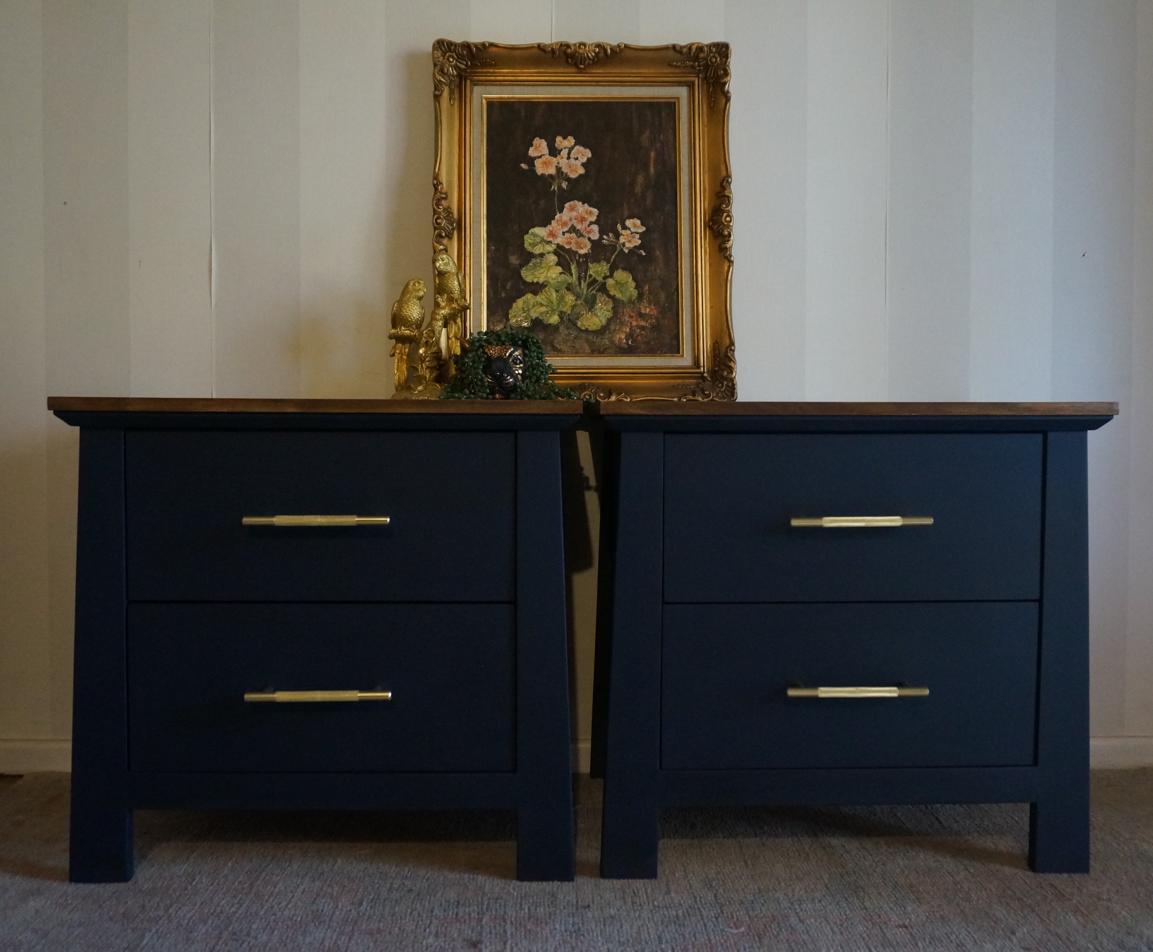 A pair of dark navy blue nightstands with gold metal handles, shown side-by-side against a white paneled wall with a framed floral painting on top.
