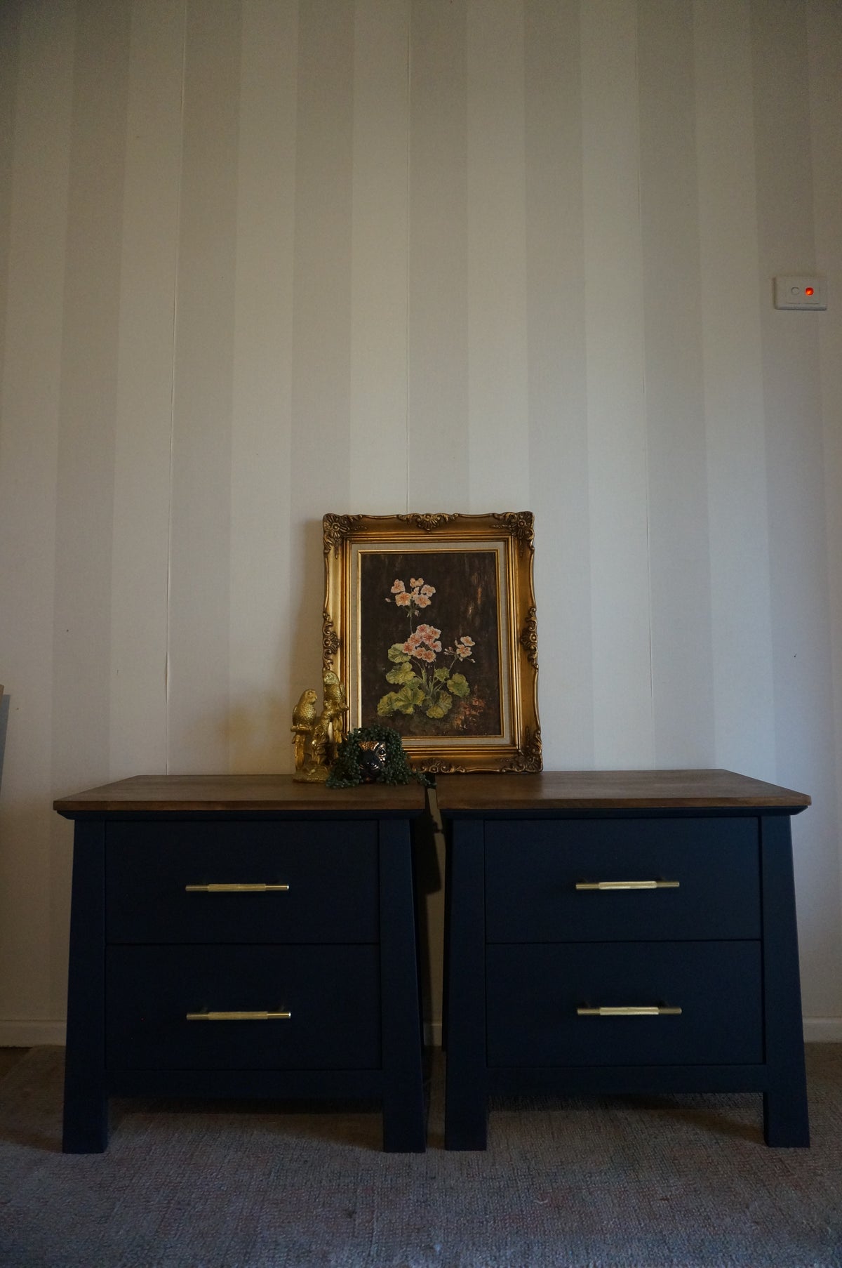 A pair of dark navy blue bedside tables with a brown wood top, featuring gold metal drawer pulls, set against a white and beige vertically striped wall with a framed floral painting on top.
