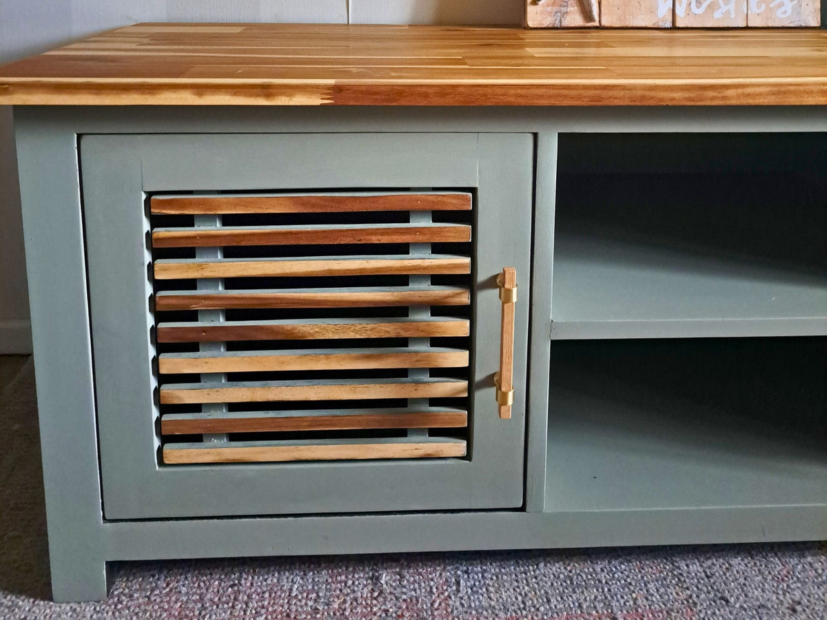 A close-up of a painted TV stand with a sage green body, a natural wood top, a slatted front panel, and open shelving in the middle.