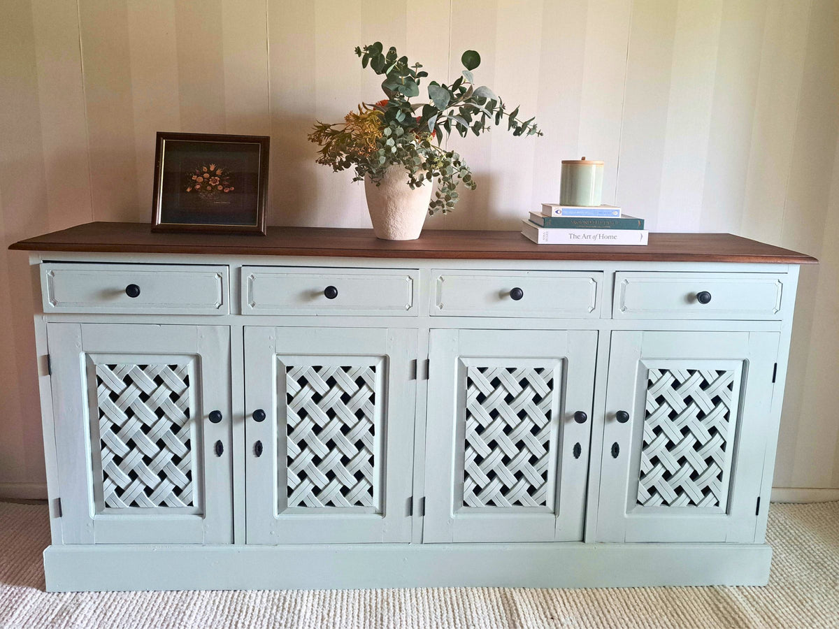 A rustic sideboard cabinet painted in Willow with a dark brown wooden top, featuring four lattice-patterned doors and styled with a vase of eucalyptus, a candle, and books on a striped wall.
