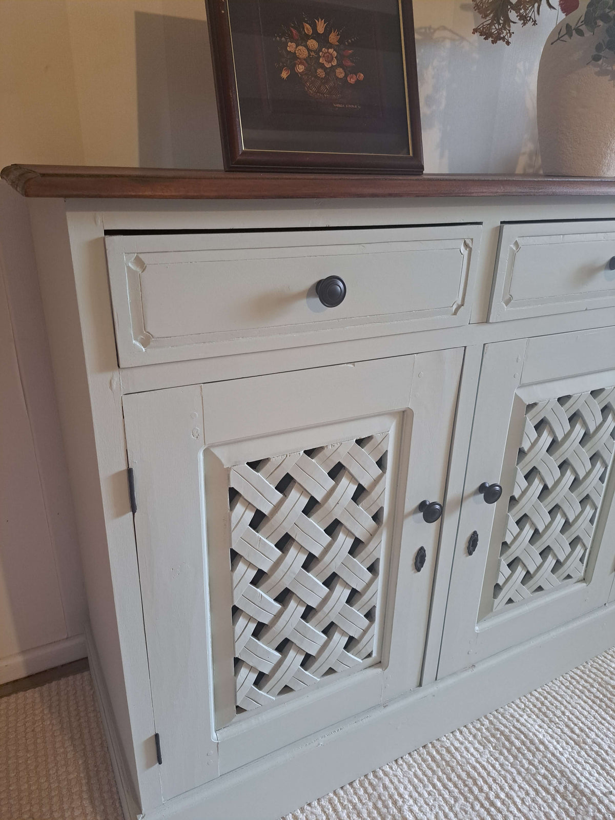 A painted wooden sideboard cabinet with a dark wood top, decorative drawer pulls, and lattice-patterned door panels, shown in a home interior setting.