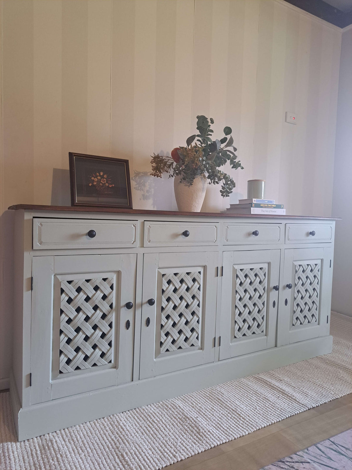 A tall, wooden sideboard cabinet with decorative lattice-patterned doors and a dark wood top, styled in a room with striped wallpaper, a woven rug, and home decor items on top.