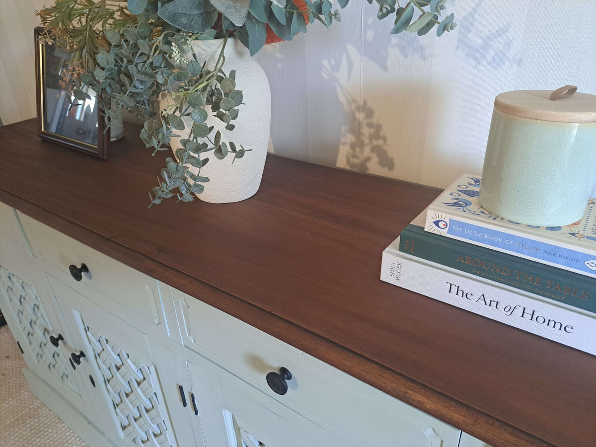 A close-up of a Willow wooden cabinet with a dark brown wood top, styled with a white ceramic vase of eucalyptus, a framed photo, and a stack of books holding a light green ceramic jar.
